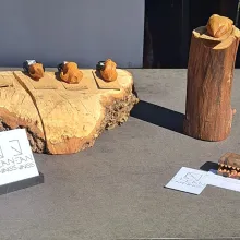 4 verschiedene aus Holz geschnitzte Schädel-Kettenanhänger liegen auf einem größeren Stück Holz. Rechts daneben steht ein dickerer Ast auf dem ein aus Holz geschnitzes anatomisches Herz liegt. Links im Vordergrund steht ein Ständer mit Visitenkarten von "clean.carvings".