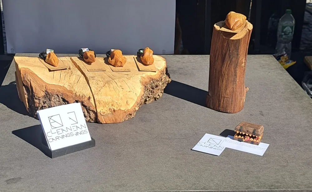4 verschiedene aus Holz geschnitzte Schädel-Kettenanhänger liegen auf einem größeren Stück Holz. Rechts daneben steht ein dickerer Ast auf dem ein aus Holz geschnitzes anatomisches Herz liegt. Links im Vordergrund steht ein Ständer mit Visitenkarten von "clean.carvings".