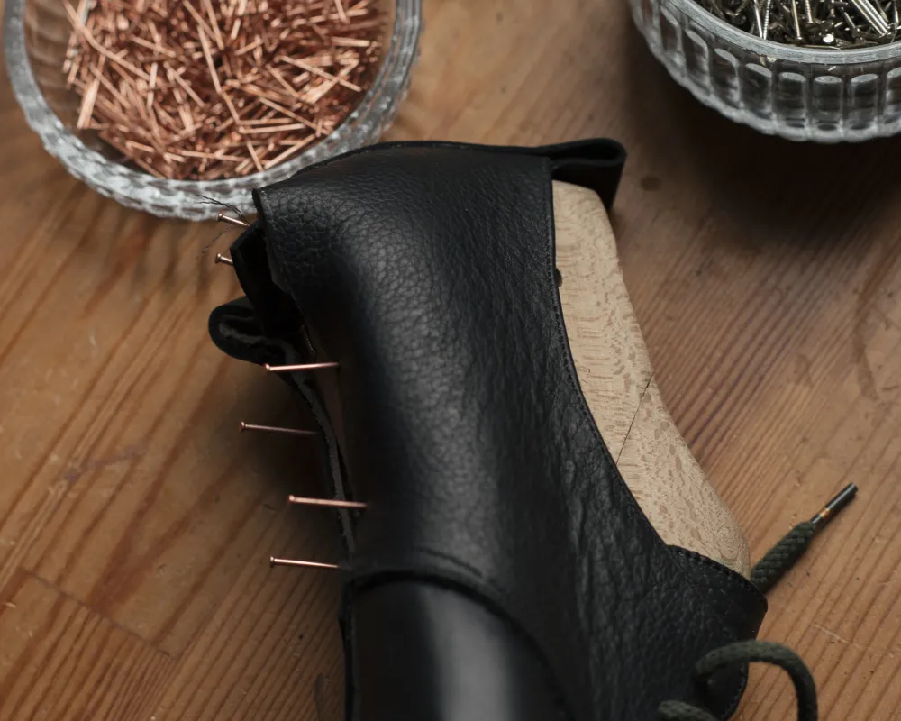 schwarze Maßschuhe liegen auf einem Tisch neben kupferfarbenen Nägeln