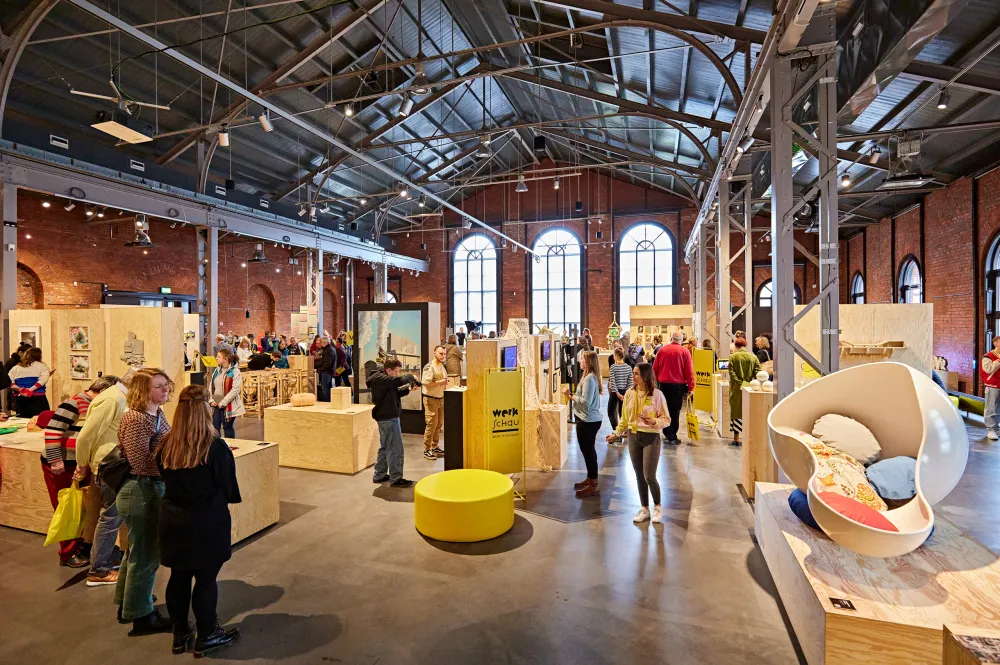 Jan Felber Eine Industriehalle mit Holzblöcken und Ausstellungsstücken, dazwischen Menschen und ein Schild mit der Aufschrift Werkschau Made in Sachsen