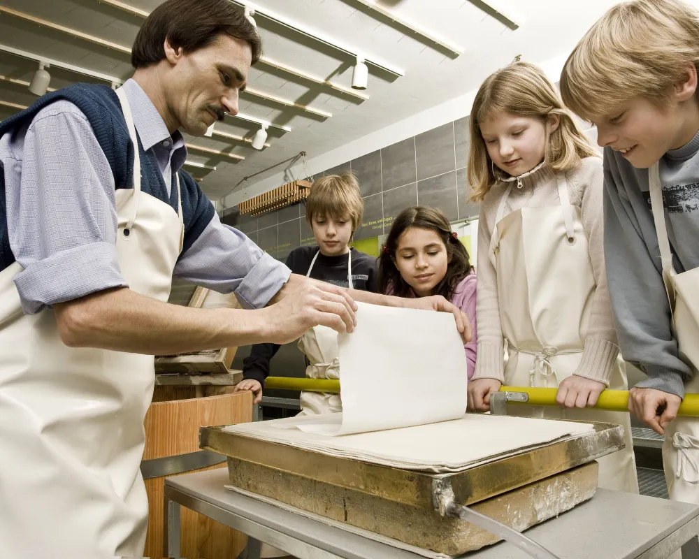 SDTB Ein Mann hält einen frisch geschöpften Bogen Papier hoch. Mehrere Kinder schauen zu.