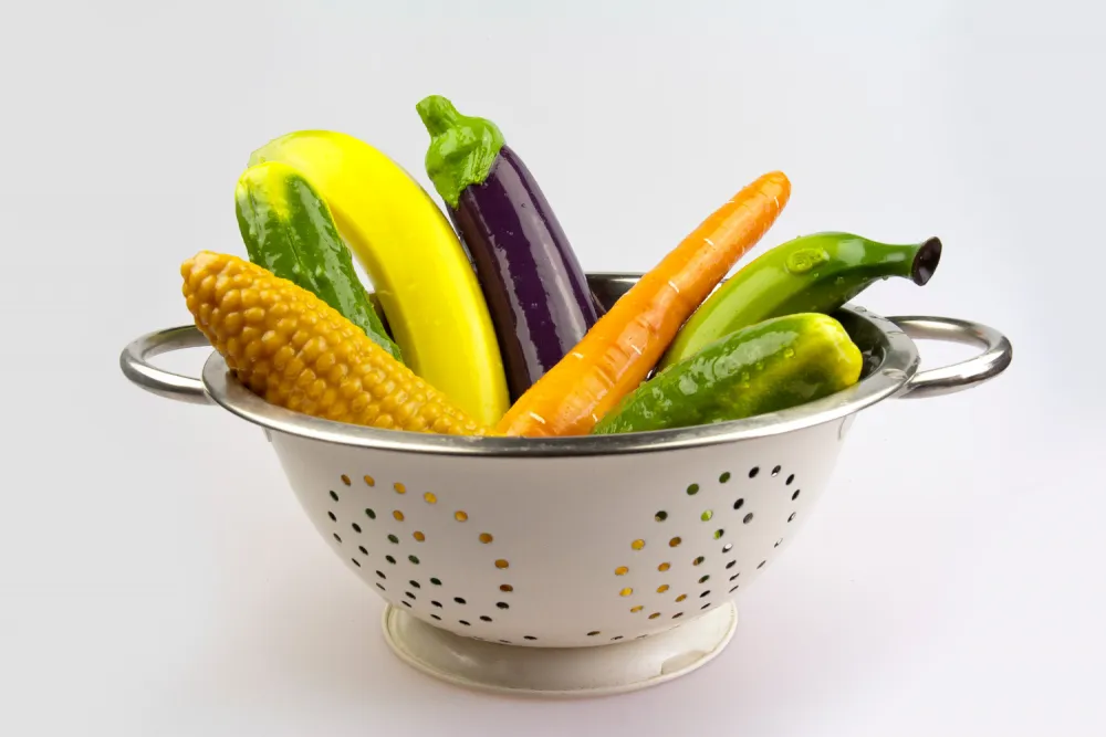 @selfdelve bunte Dildos in Form einer Banane, Aubergine, Möhre, Mais und Gurke liegen in einem Küchensieb.