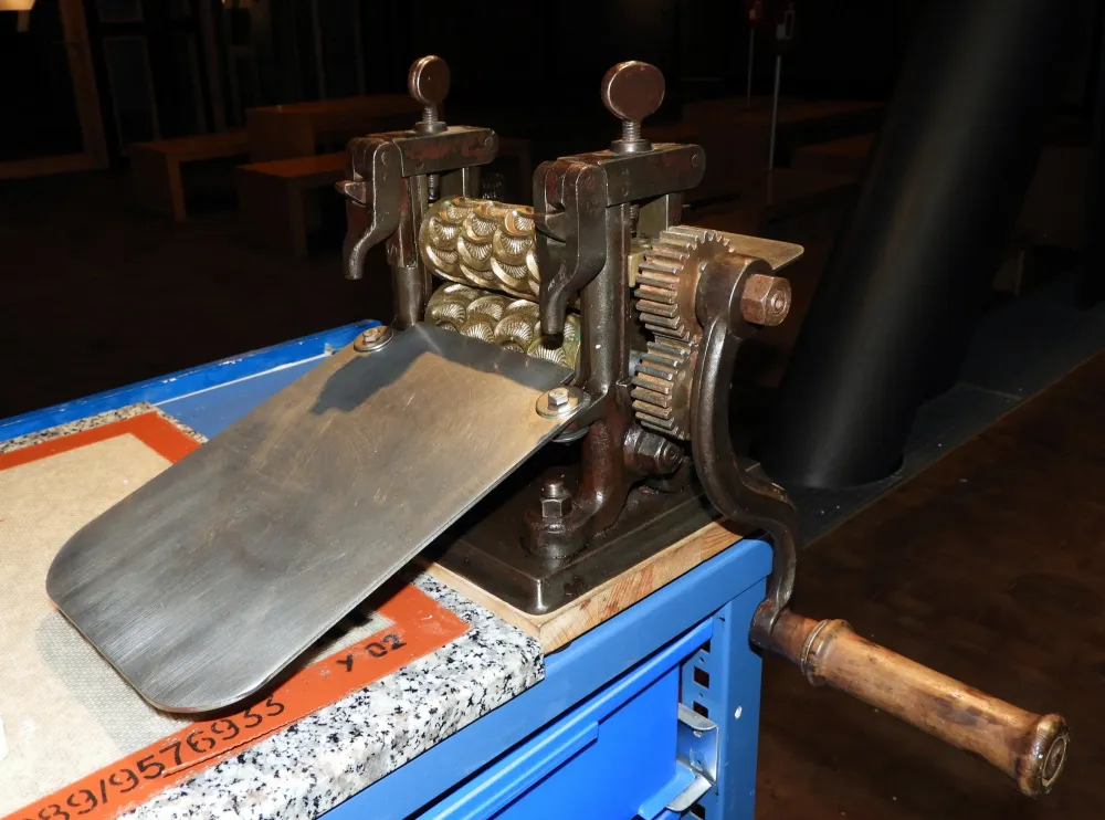 Hisorische Bonbonproduktion Man sieht eine kleine Walze. Die Metallwalzen weisen Hohlräume in Bonbonform auf. Eine Rutsche führt von den Walzen hinab auf einen Tisch.
